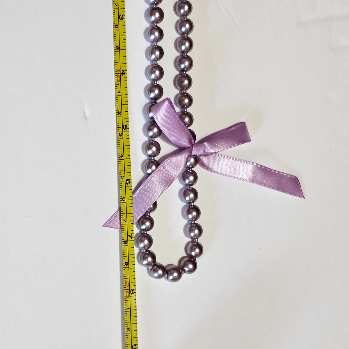 Purple beaded necklace with a ribbon bow, measured against a ruler on a white background